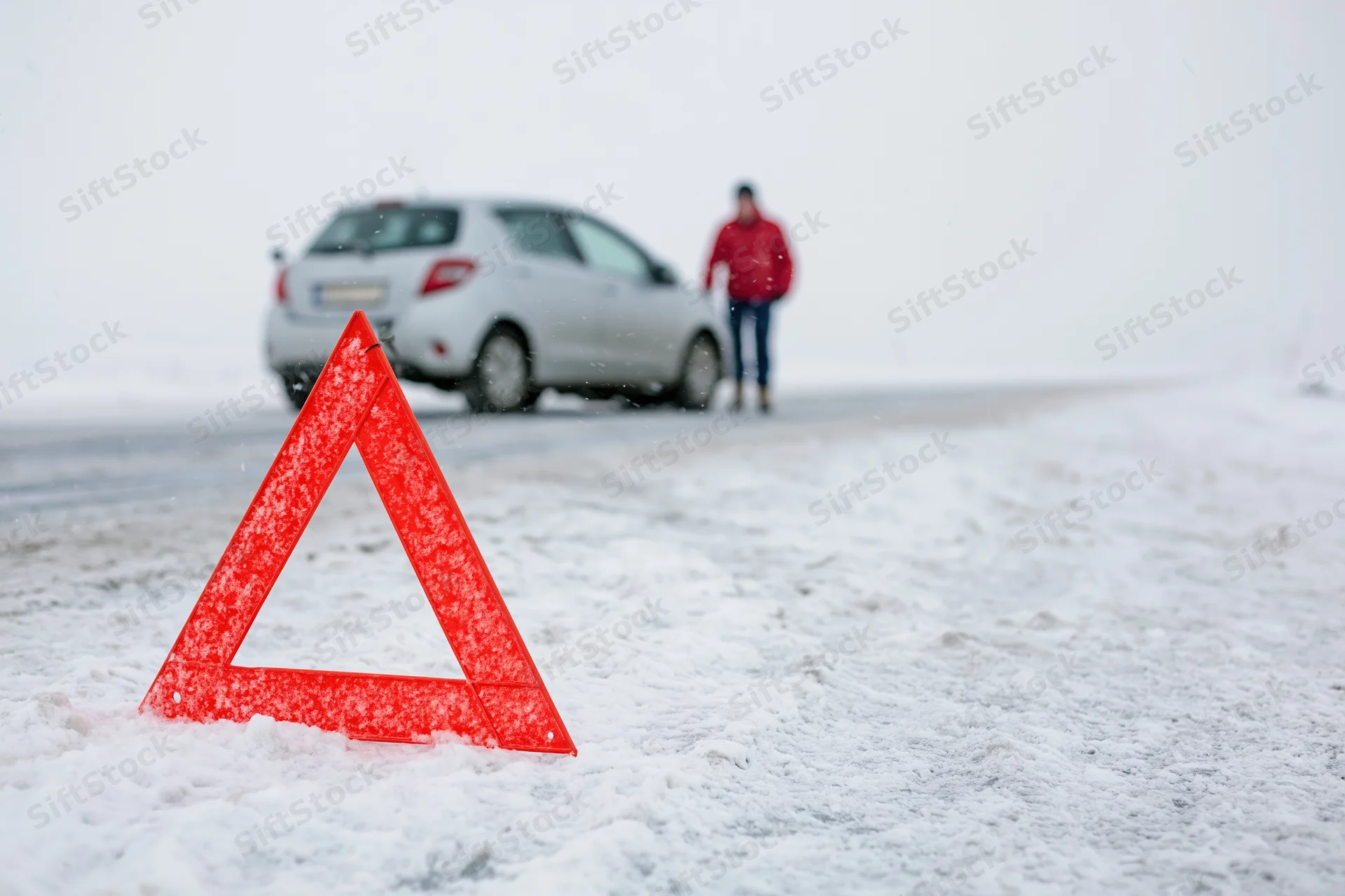 Red emergency warning triangle on snow-covered road with car and person in background