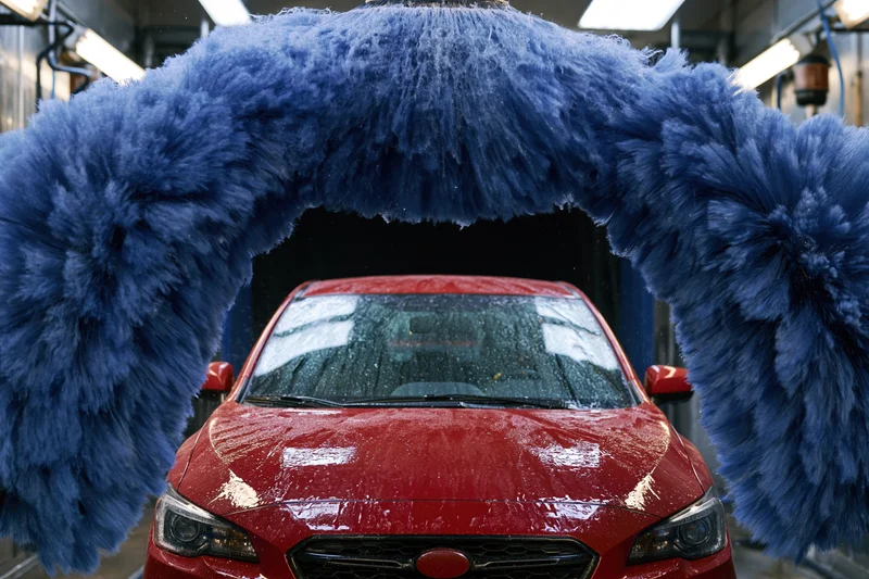 Red sedan moving through automatic car wash tunnel flanked by large blue brushes