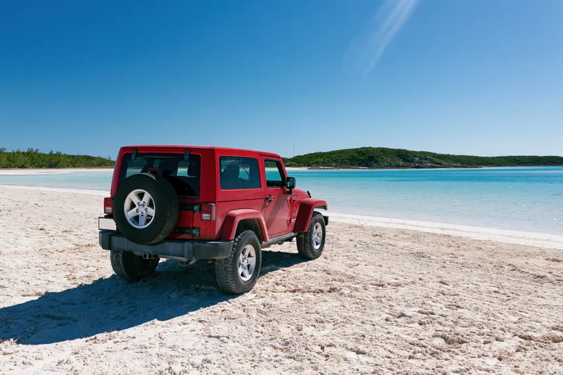 Red two-door suv parked on white sand beach facing ocean under clear blue sky