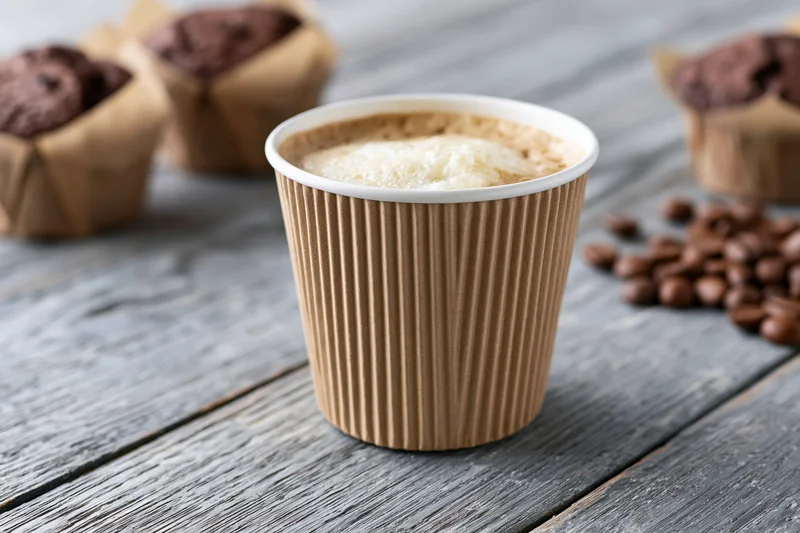 Ripple-wrap paper coffee cup filled with latte on a rustic wooden table with chocolate muffins and scattered coffee beans nearby