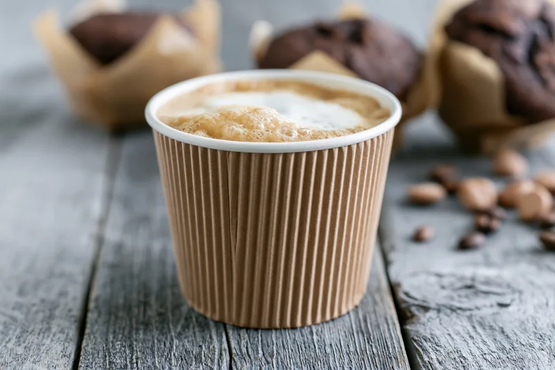Ripple-wrap paper coffee cup filled with latte foam on a dark wooden table with chocolate muffins and scattered coffee beans nearby