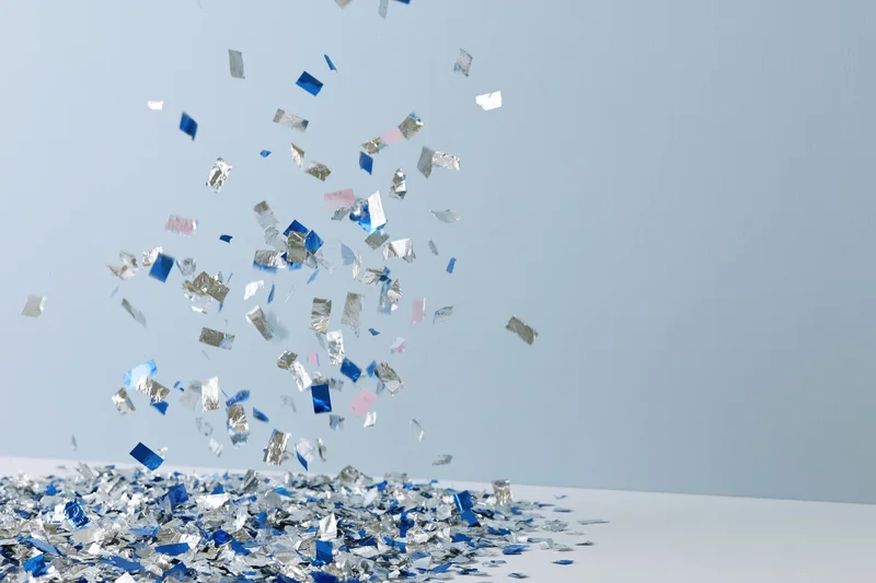 Silver and blue metallic confetti pieces falling midair against a light blue background with a pile on the surface below