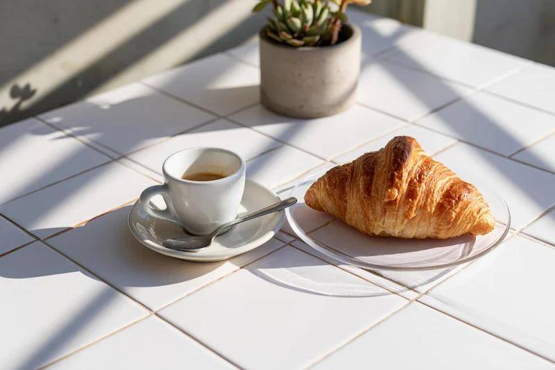 Espresso cup with spoon on saucer and croissant on plate on white tiled table in sunlight