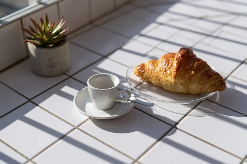 Croissant and espresso cup on tiled windowsill with sunlight and shadows