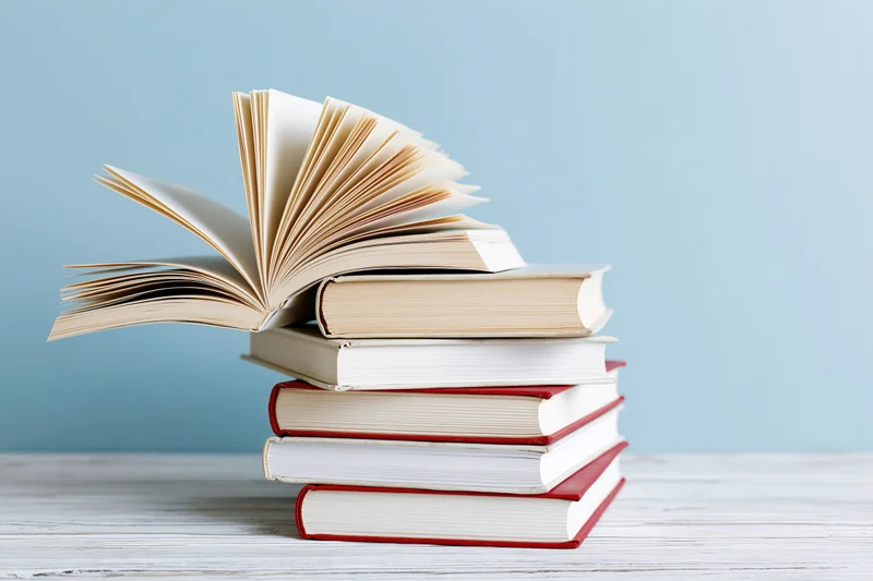 Stack of open hardcover books piled on a white wooden surface with blue background