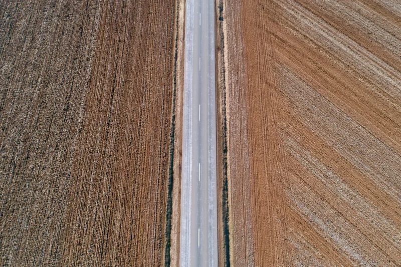 Straight two-lane asphalt road cutting through plowed farmland from above