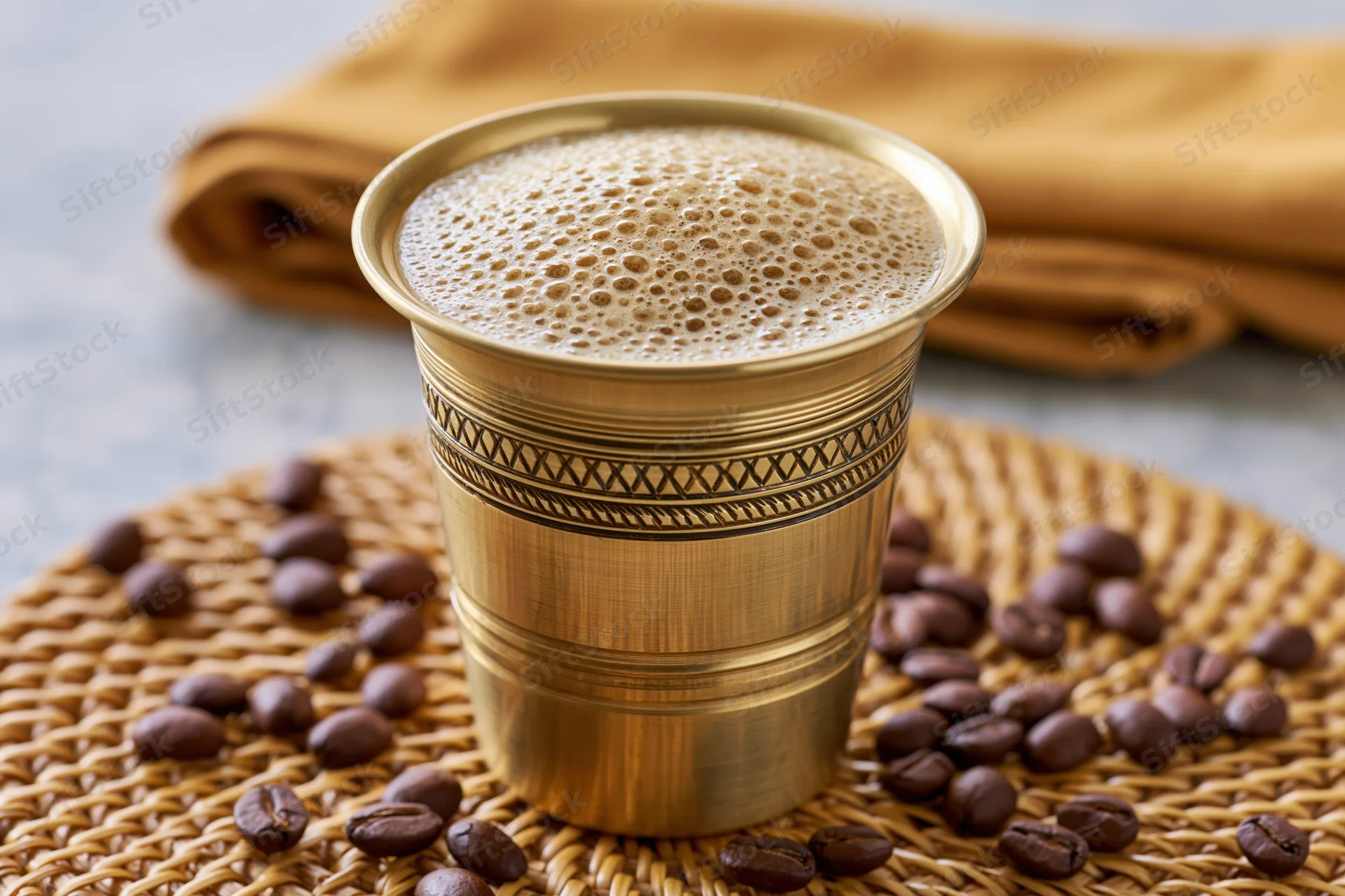 Traditional south indian filter coffee in a brass tumbler with froth and coffee beans on a woven placemat