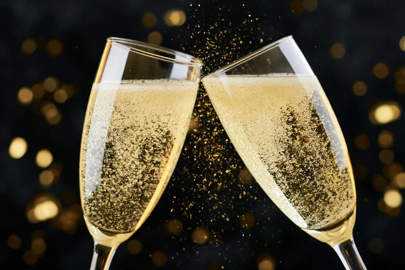 Two champagne flutes clinking together with golden bubbles and glitter against a dark bokeh background