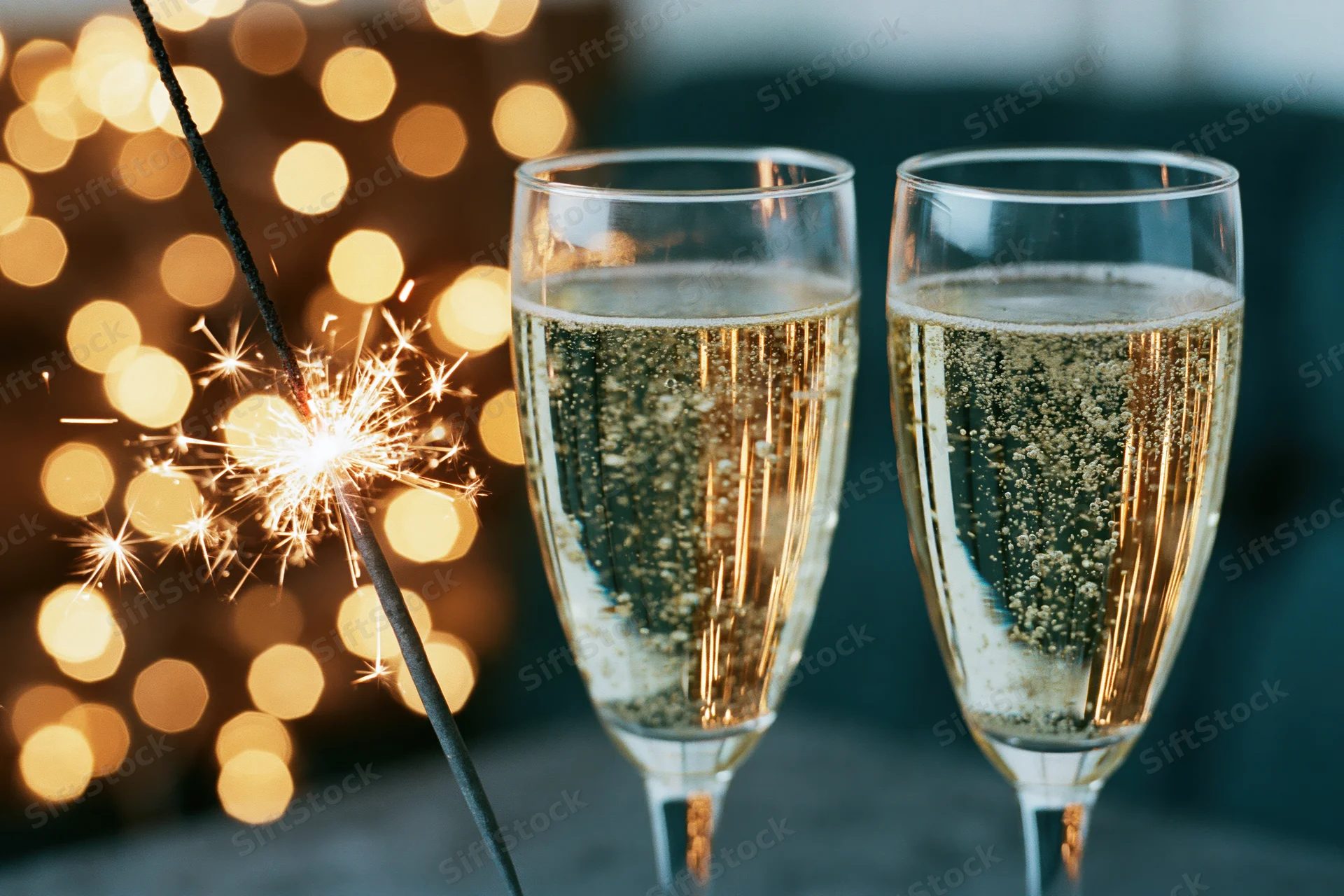 Two champagne flutes filled with sparkling wine beside a lit sparkler on a table with bokeh lights in the background