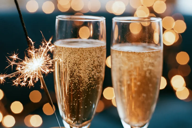 Two champagne flutes filled with sparkling wine beside a lit sparkler against warm bokeh lights
