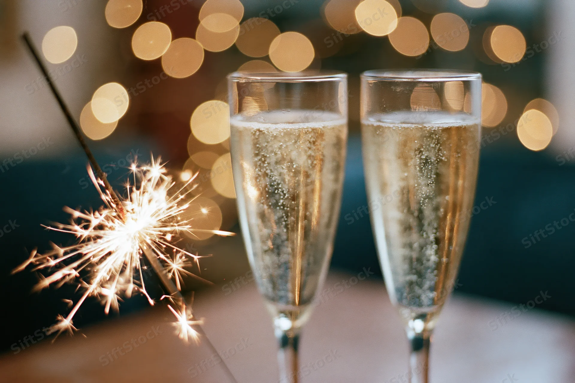 Two champagne flutes with sparkling wine and a lit sparkler on a table with bokeh lights in the background
