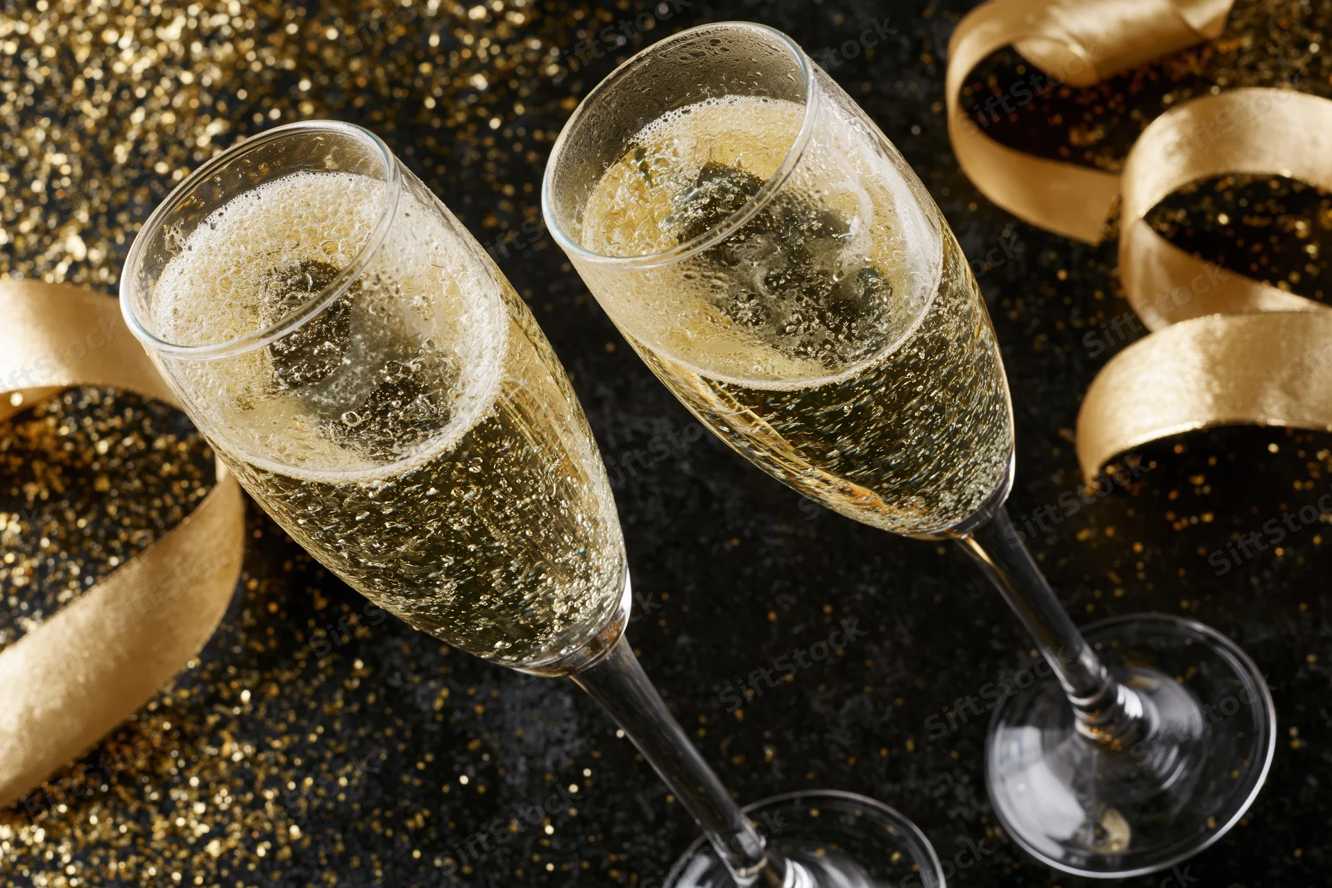 Two champagne flutes clinking together on a glittering black surface with golden ribbons