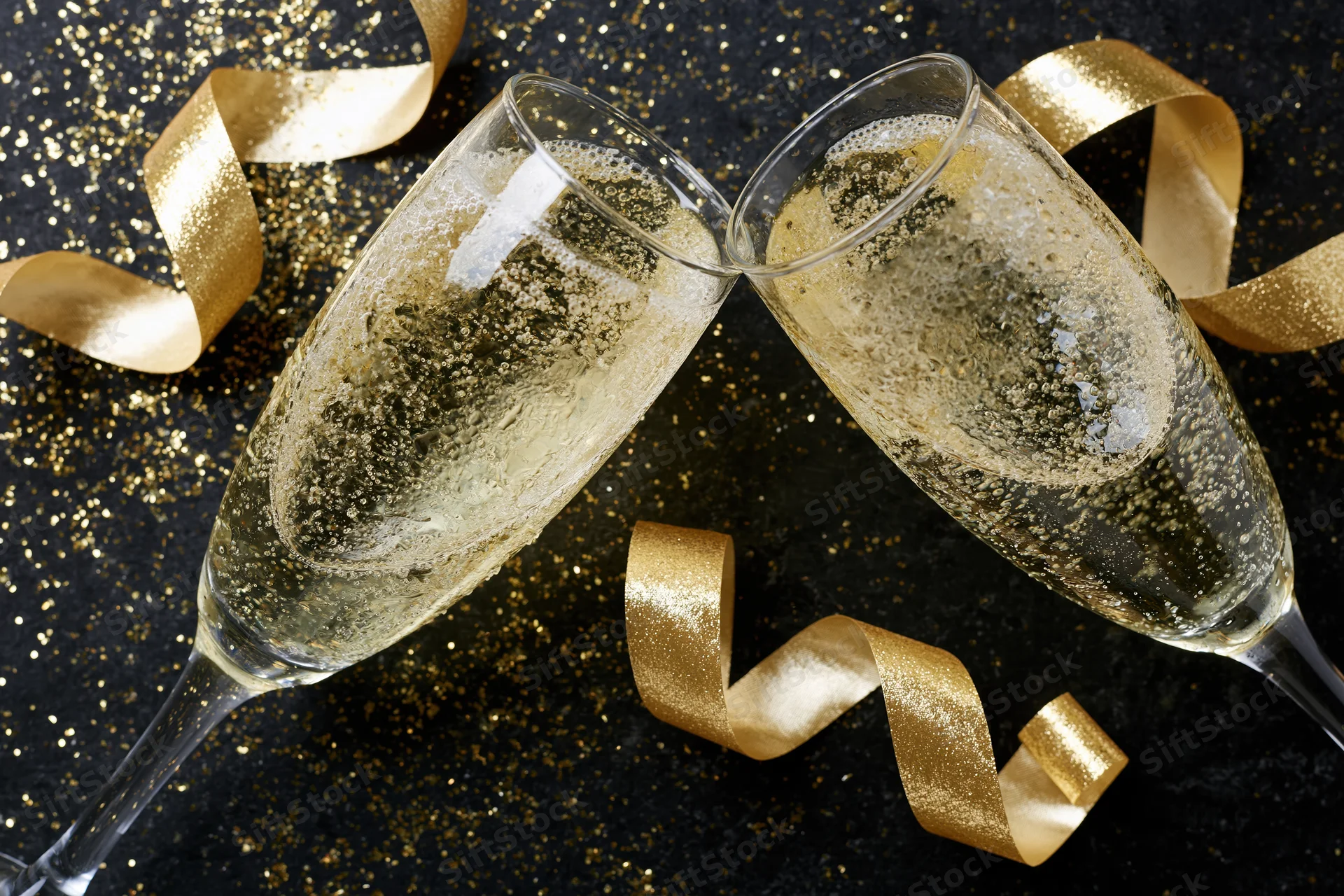Two champagne flutes clinking together on a black glittery surface with golden ribbons scattered around