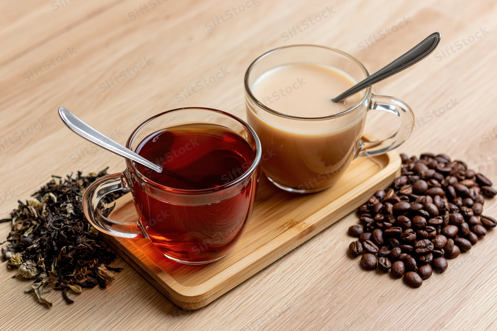 Two glass mugs with tea and coffee on a bamboo tray beside loose tea leaves and coffee beans on a wooden table