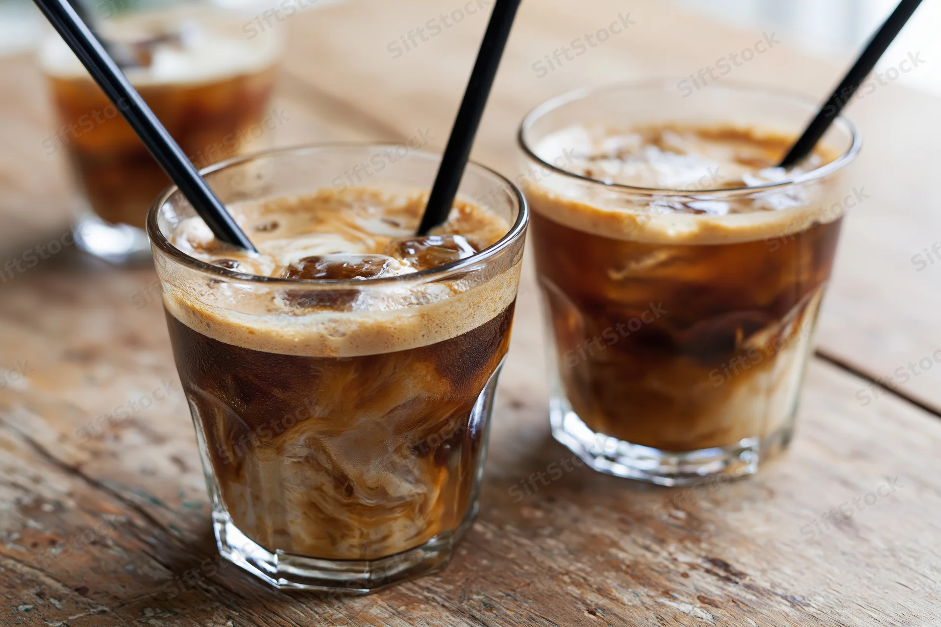 Three short glasses of iced coffee with cream swirls and black straws on a rustic wooden table
