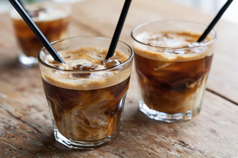 Three short glasses of iced coffee with cream swirls and black straws on a rustic wooden table