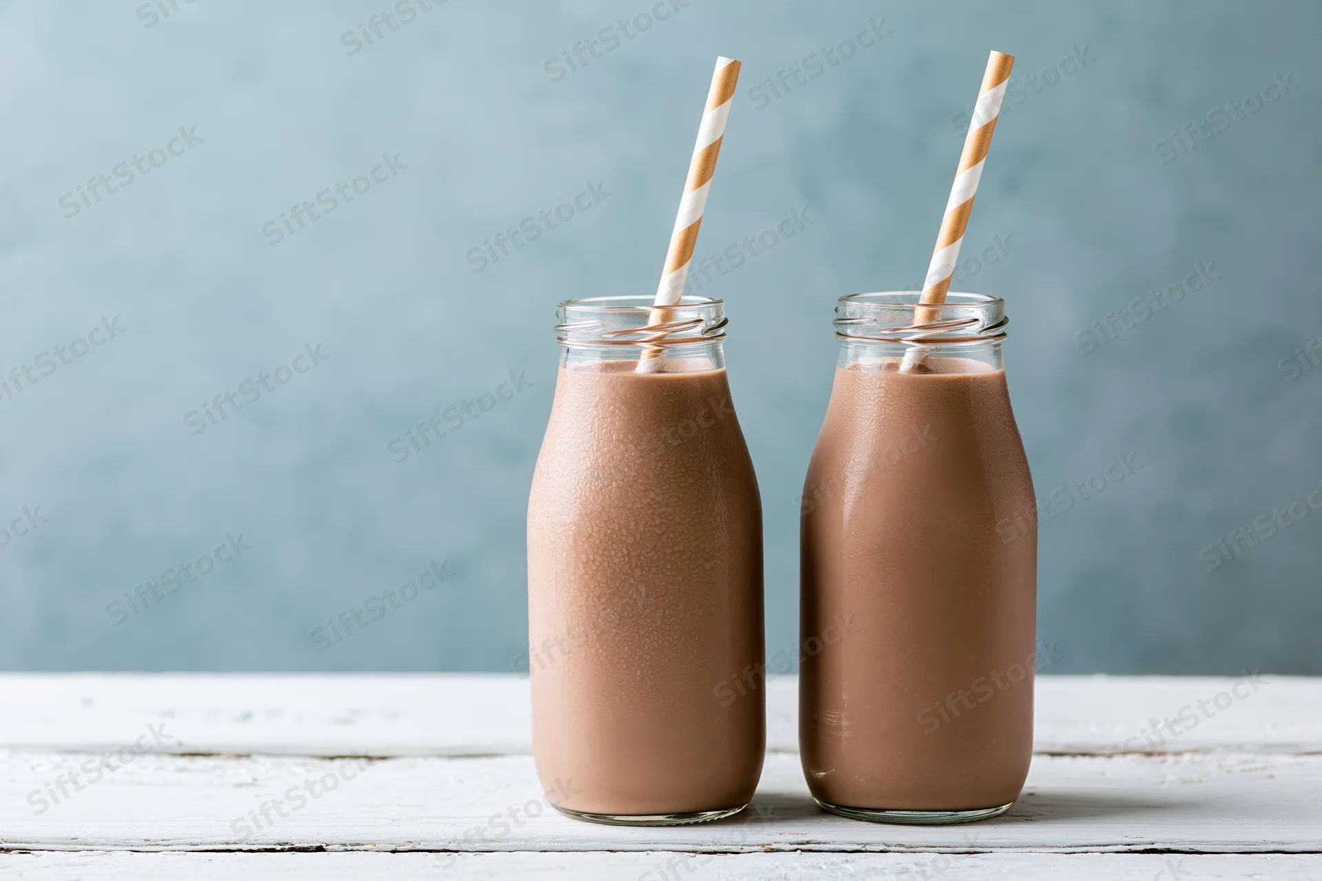 Two glass bottles of chocolate milk with striped straws on a white wooden table