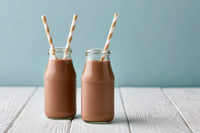 Two glass bottles filled with chocolate milk on a white wooden table, each with a striped paper straw