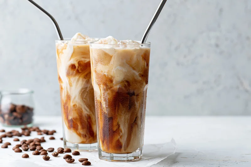 Two tall glasses of iced coffee with cream swirls and metal straws on a white surface with scattered coffee beans