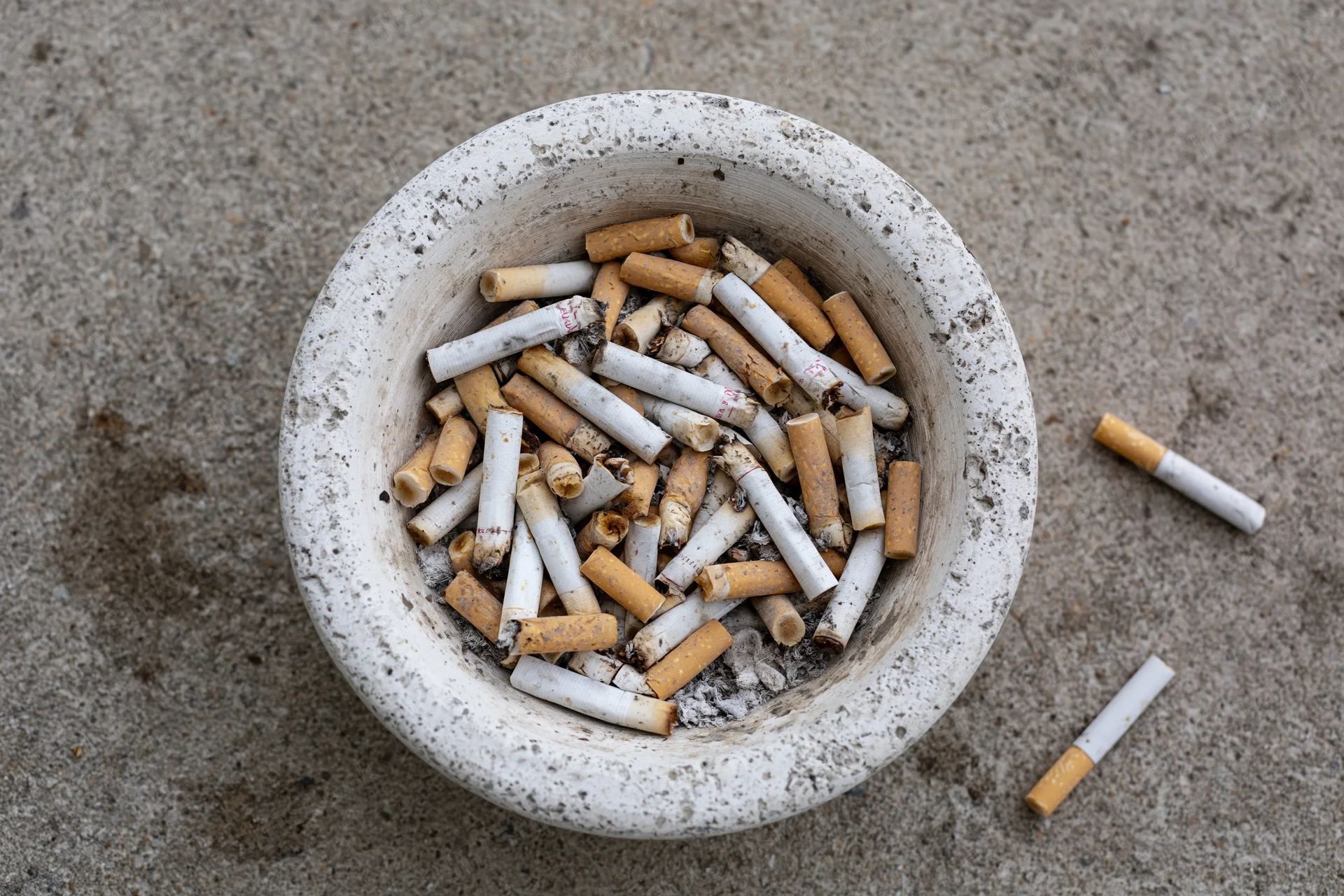 Weathered white concrete ashtray overflowing with cigarette butts on sandy ground