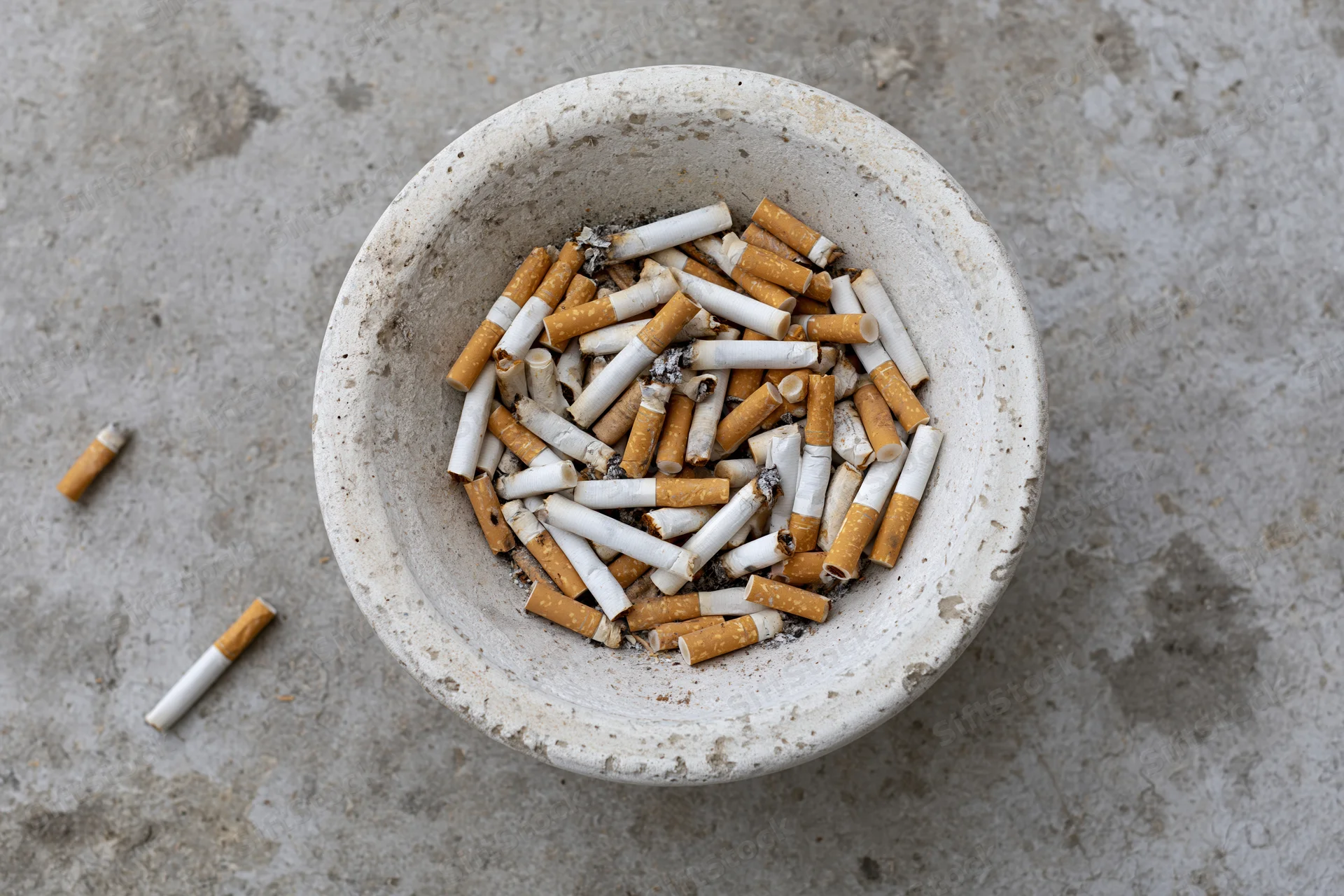 Weathered white concrete ashtray overflowing with cigarette butts on a gray textured surface
