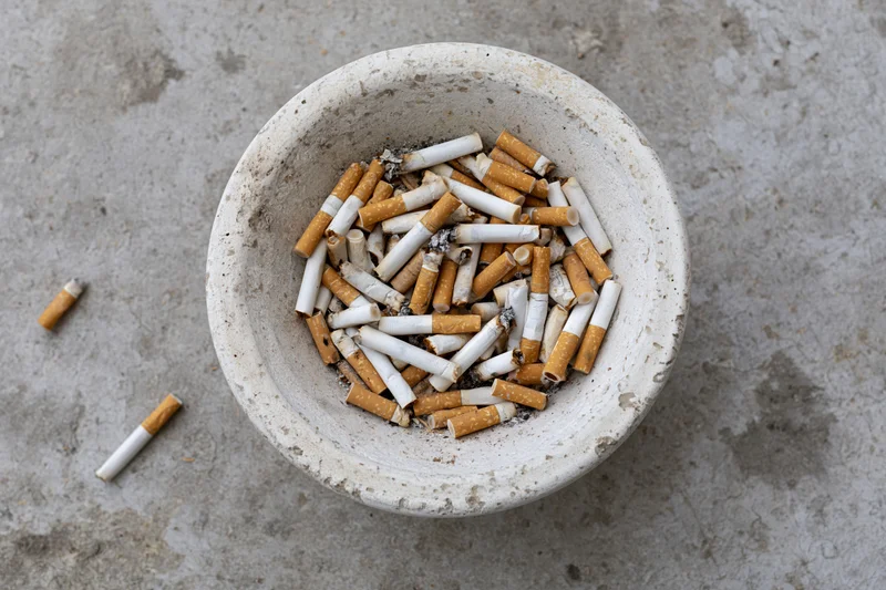 Weathered white concrete ashtray overflowing with cigarette butts on a gray textured surface