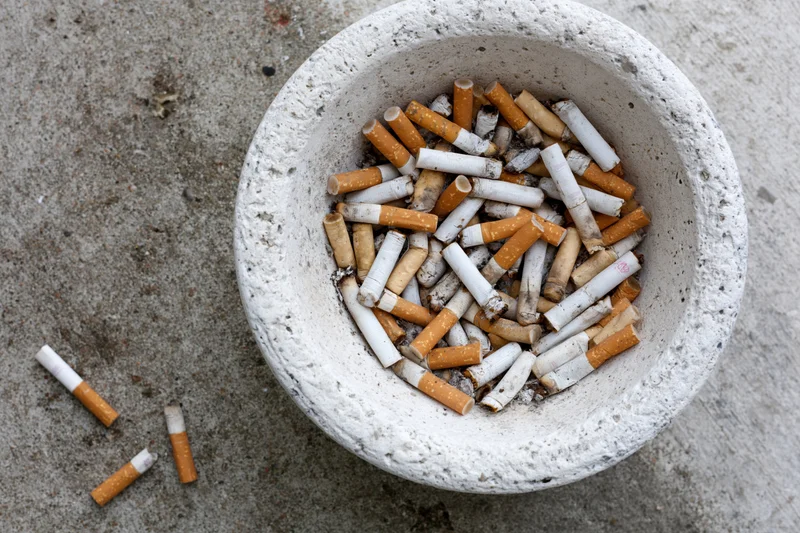 Weathered white concrete ashtray overflowing with used cigarette butts on gray pavement