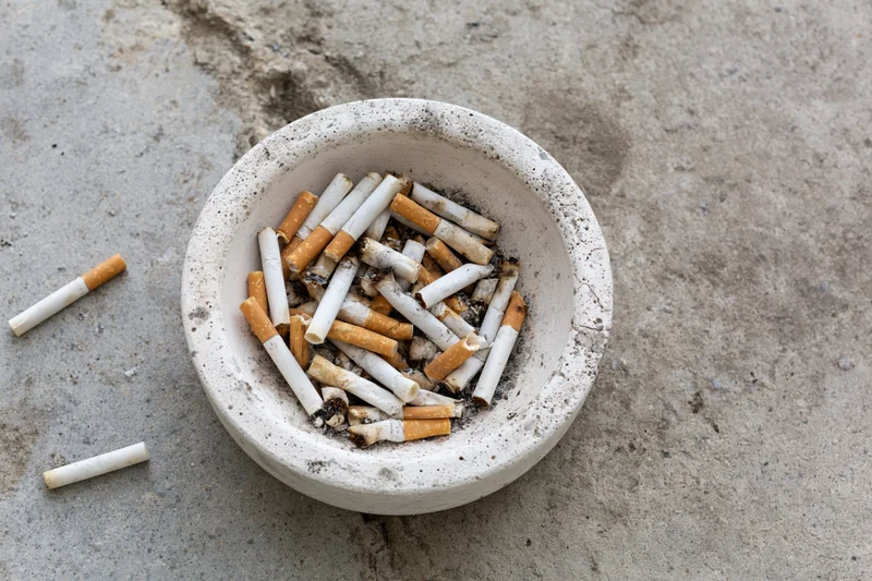 Weathered white concrete ashtray overflowing with cigarette butts on a gray pavement