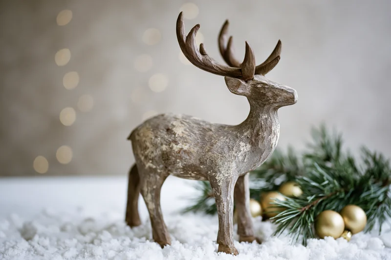 Weathered wooden reindeer figurine standing on snow beside pine branches and gold ornaments with bokeh lights in background