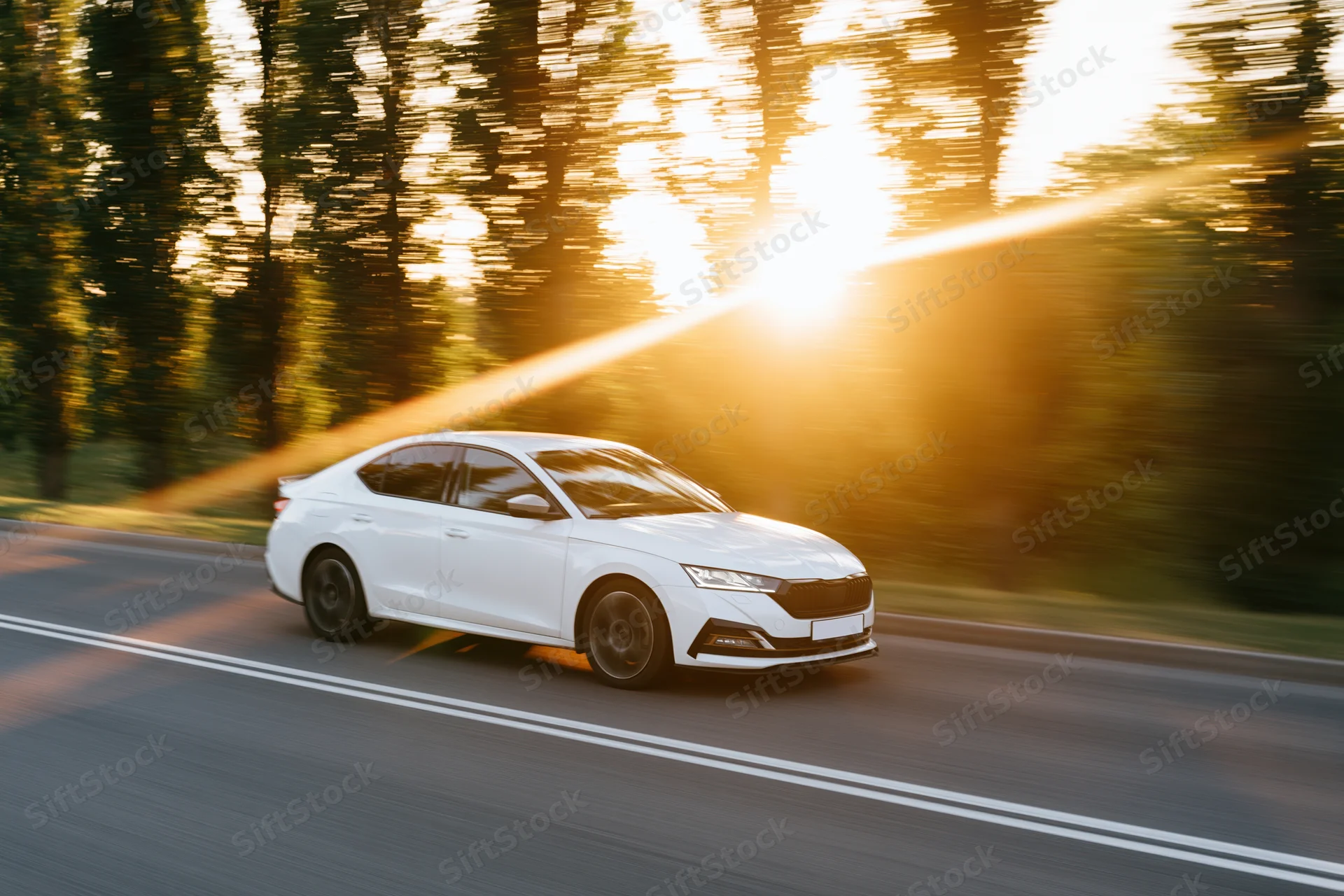 White sedan driving at speed on a two-lane road at sunset with trees in motion blur