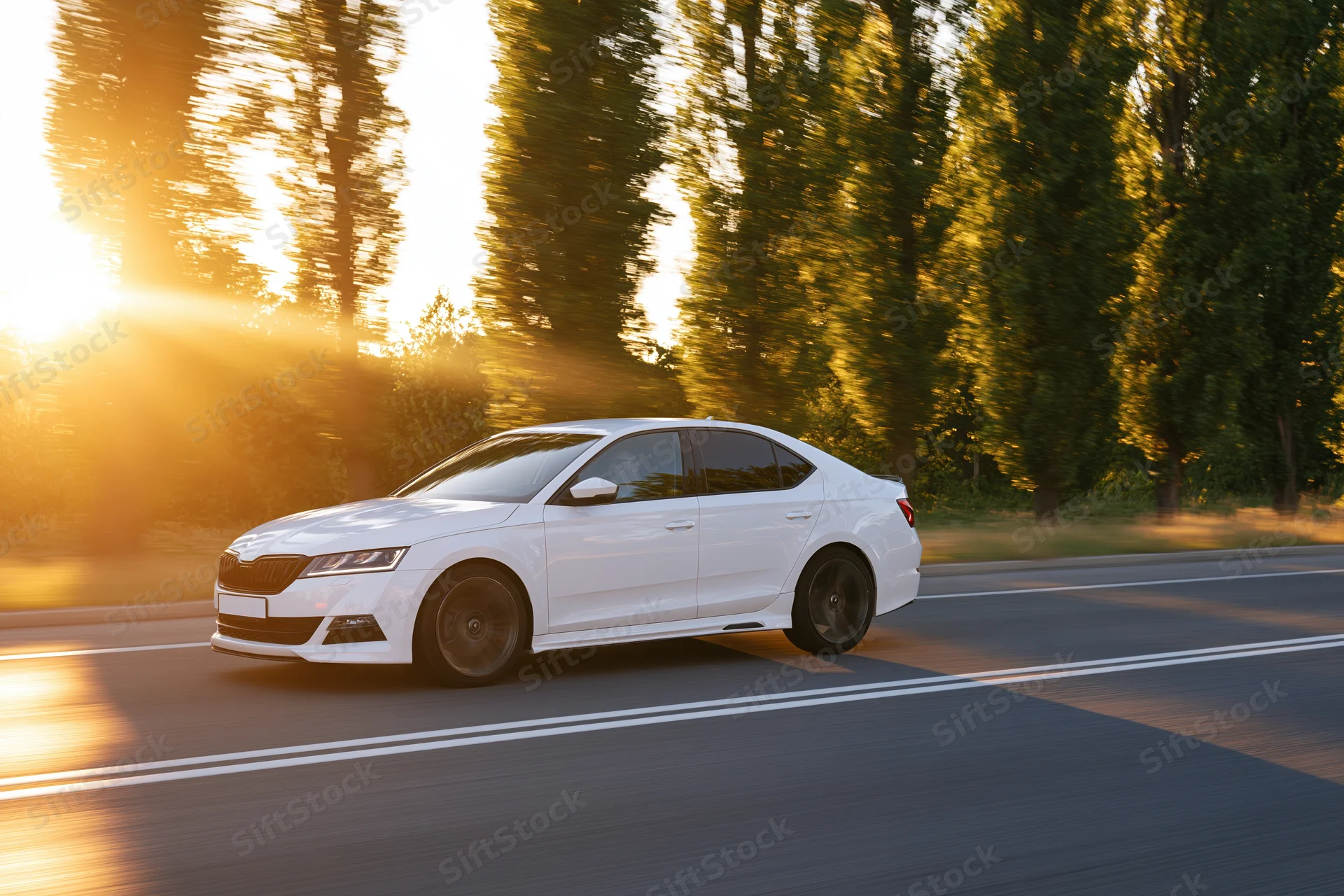 White sedan driving at speed on a two-lane road lined with trees at sunset