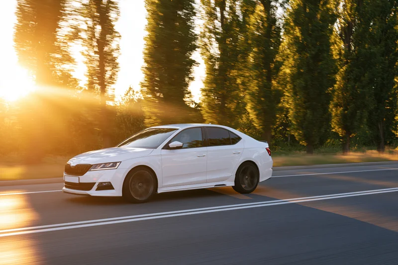 White sedan driving at speed on a two-lane road lined with trees at sunset