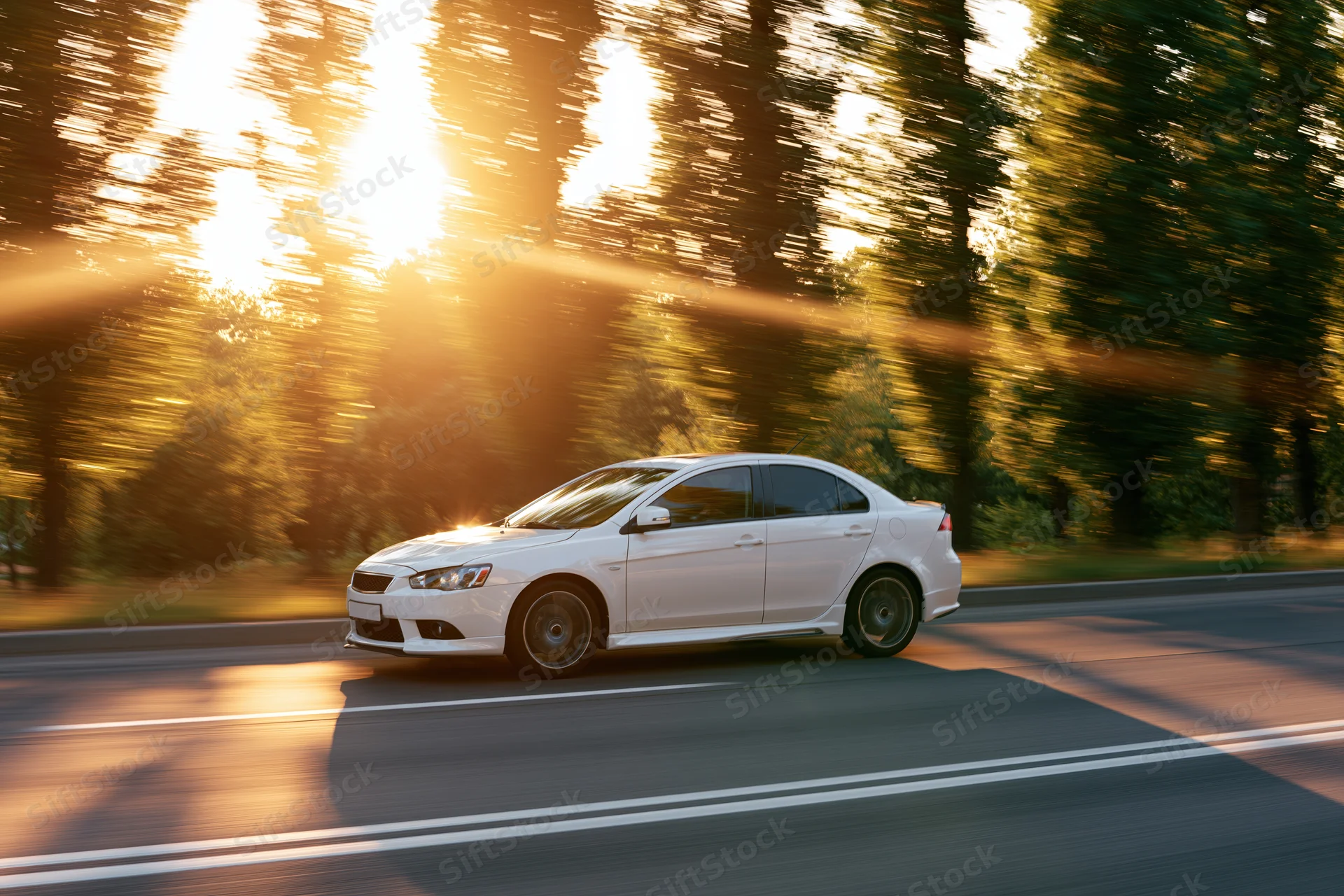 White sport sedan driving at speed on a two-lane road lined with trees at sunset
