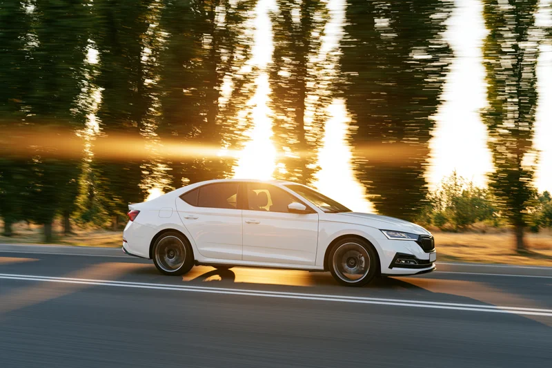 White sedan driving at speed on a two-lane road lined with trees at sunset