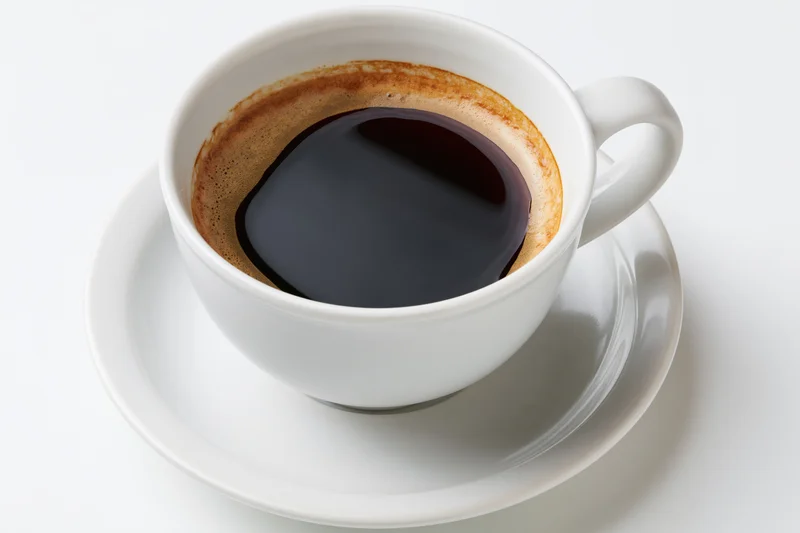White ceramic coffee cup filled with black coffee and crema on a saucer against a white background