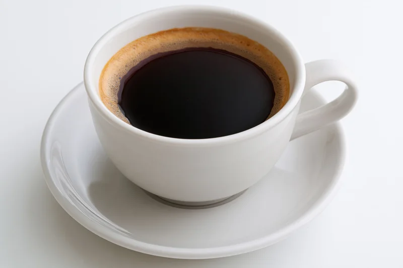 White ceramic coffee cup filled with black coffee and crema on a saucer against a white background
