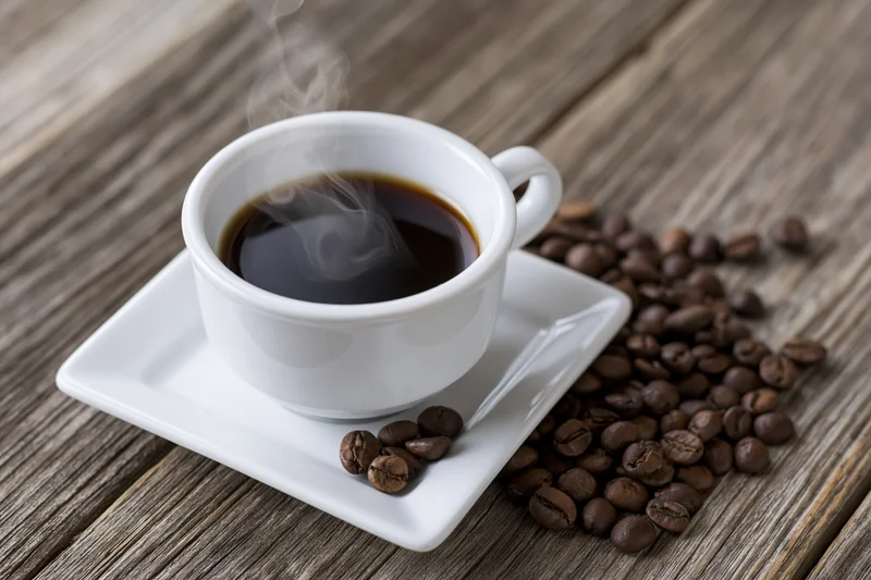 Steaming coffee in a white ceramic cup on a square saucer with roasted beans scattered on a wooden table