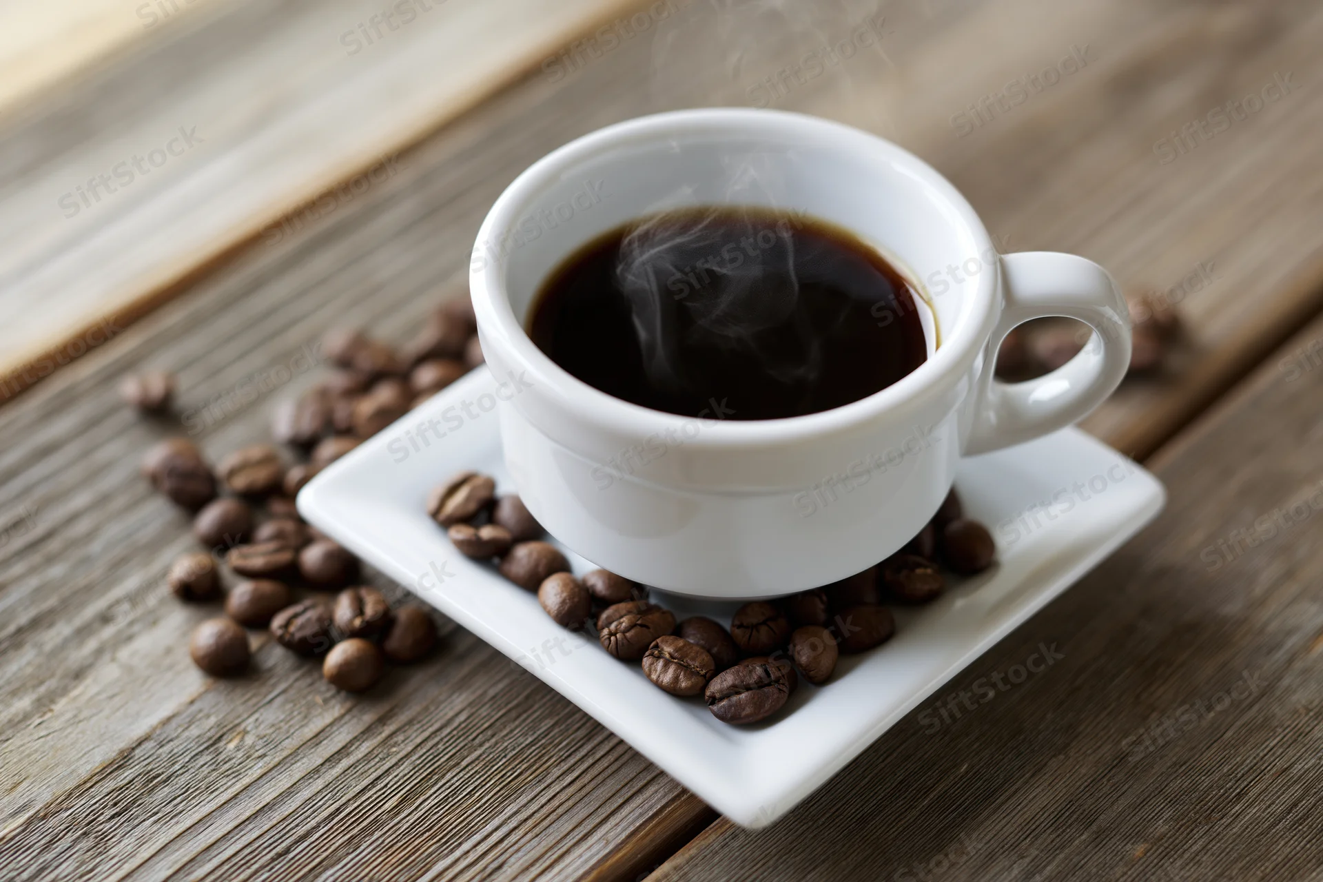 Steaming coffee in a white ceramic cup on a square saucer with scattered beans on a wooden table