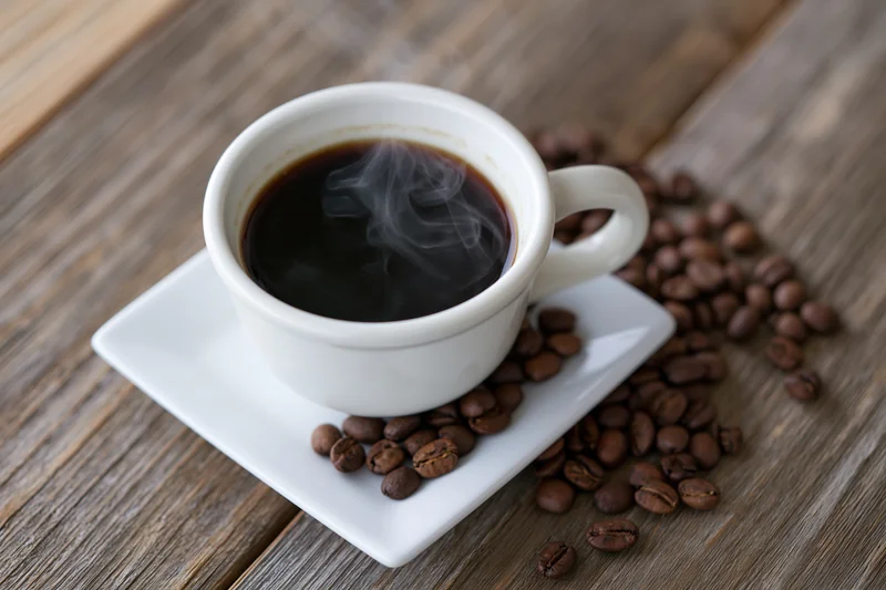 Steaming coffee in a white ceramic cup on a square saucer with scattered beans on a wooden table