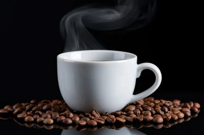 White ceramic coffee cup with rising steam resting on roasted coffee beans on a reflective black surface