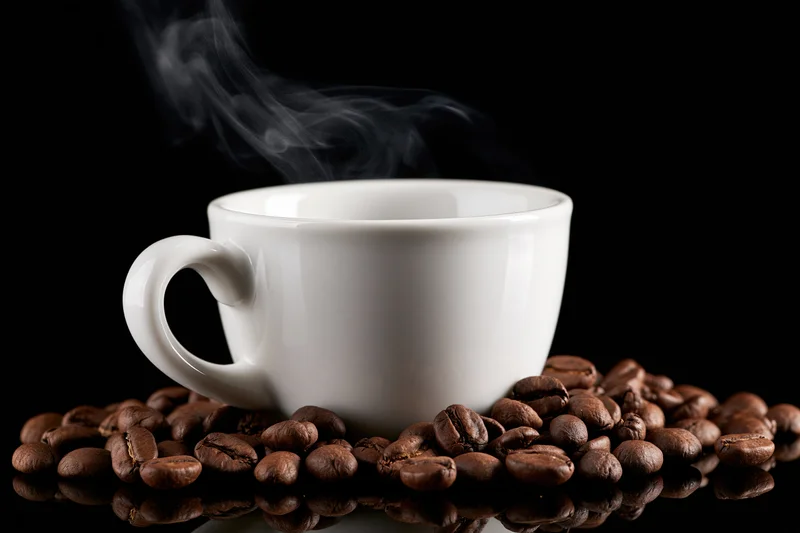 White ceramic coffee cup steaming on a bed of roasted coffee beans against black background