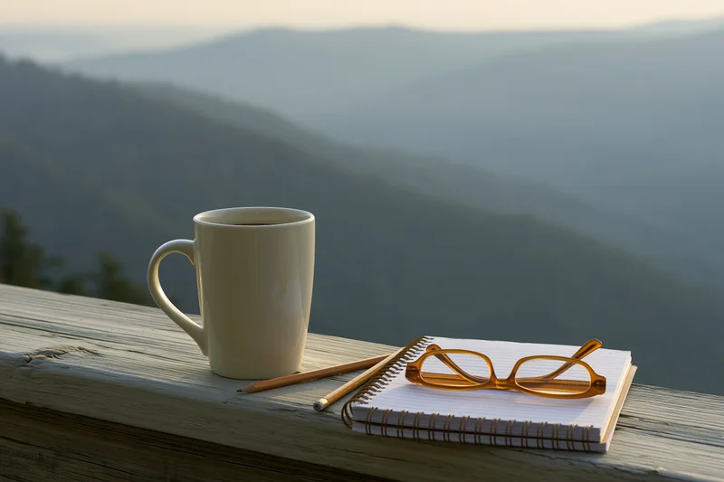 White ceramic coffee mug with notebook, pencil and glasses on wooden railing overlooking mountain landscape at sunrise