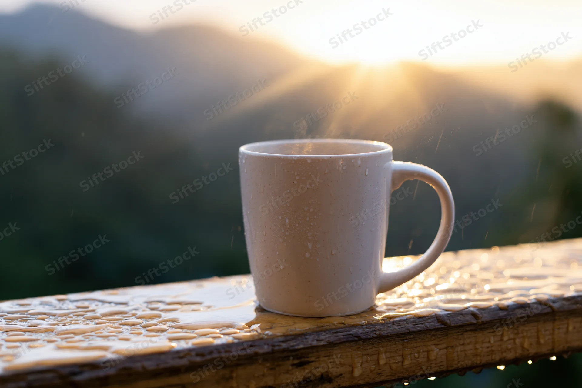 White ceramic coffee mug steaming on wet wooden railing at sunrise overlooking mountains and forest
