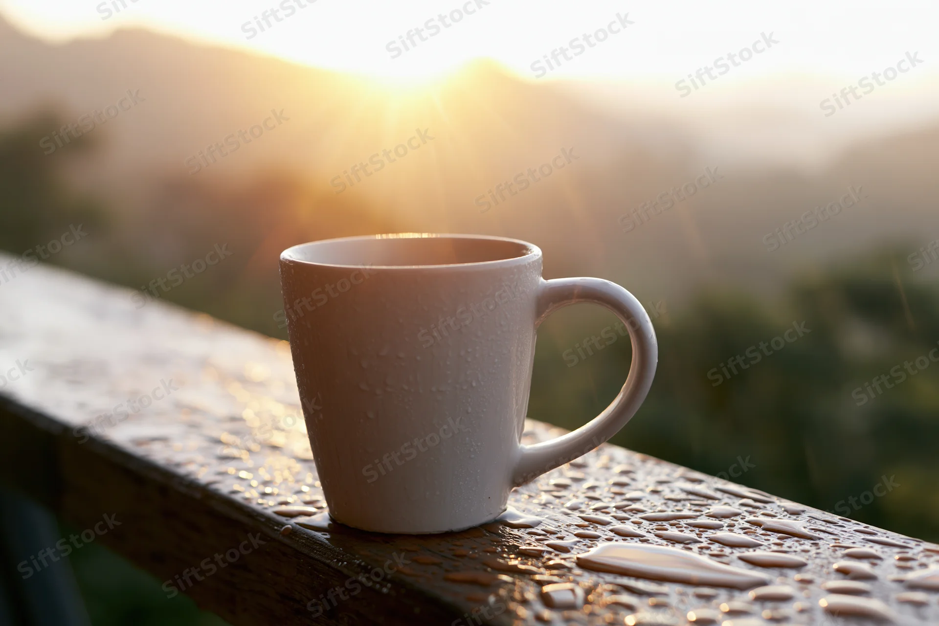 White ceramic coffee mug with water droplets on wet wooden railing at sunrise overlooking mountain landscape