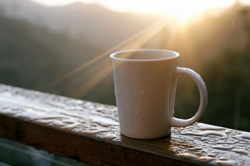 White ceramic coffee mug with water droplets on wet wooden railing at sunrise overlooking mountains