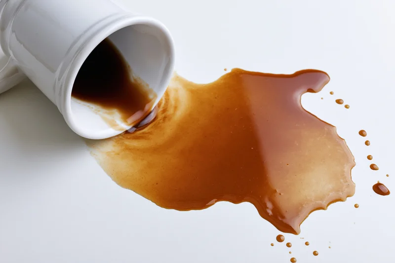 White ceramic coffee mug tipped over with dark brown liquid spilling onto a smooth white surface