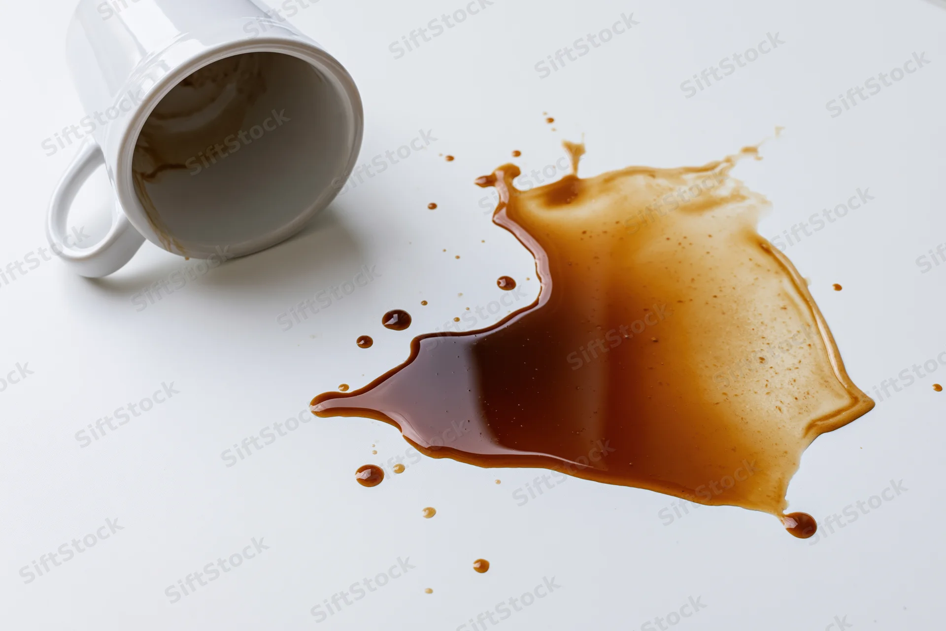 White ceramic coffee mug tipped over with dark brown liquid spilled on a white surface