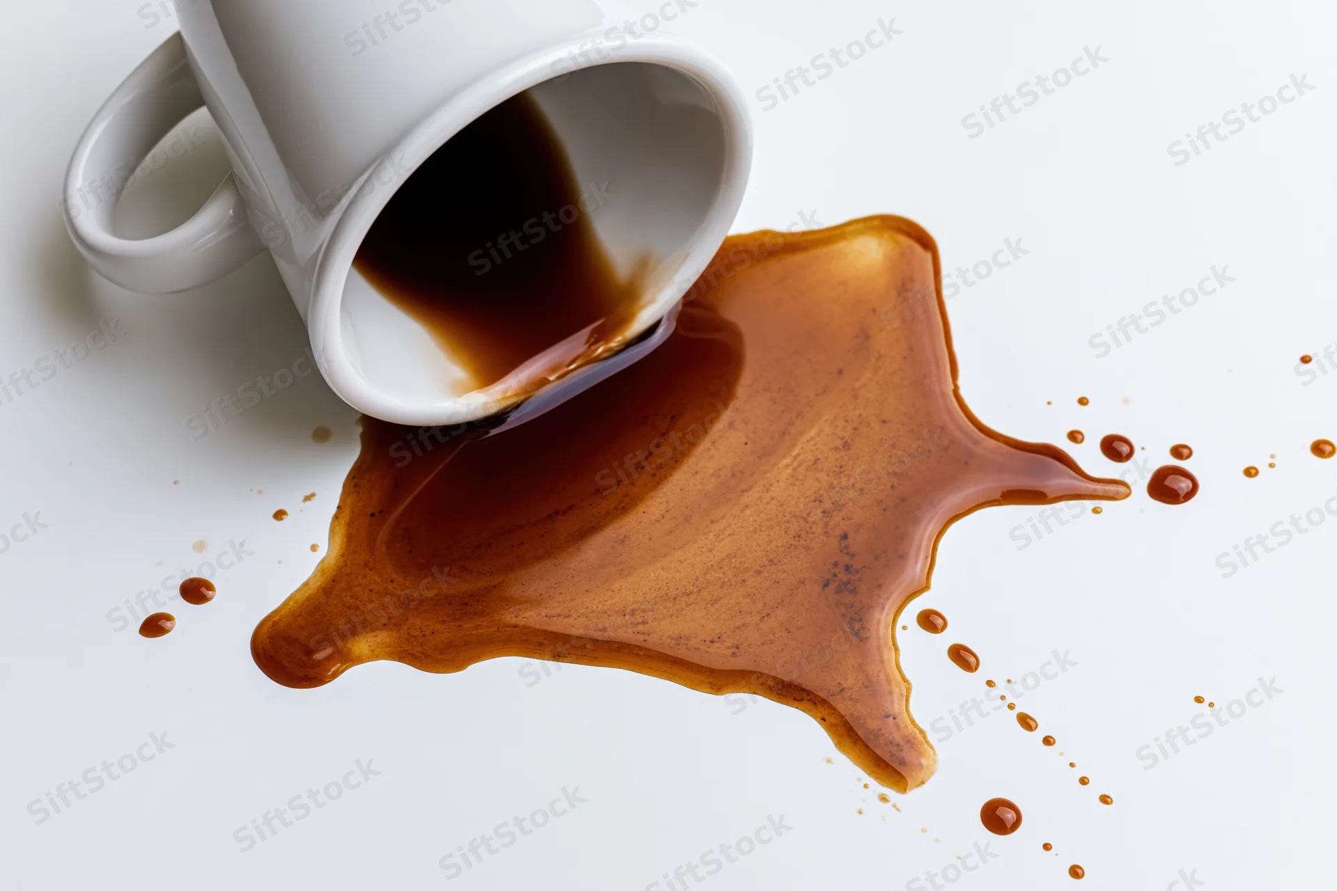 White ceramic coffee mug tipped over with dark brown liquid spilling onto a white surface