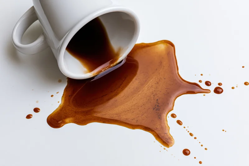 White ceramic coffee mug tipped over with dark brown liquid spilling onto a white surface