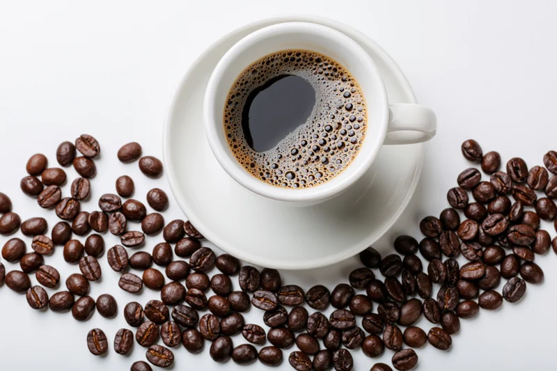 White ceramic cup of black coffee with crema on saucer surrounded by roasted coffee beans on white surface
